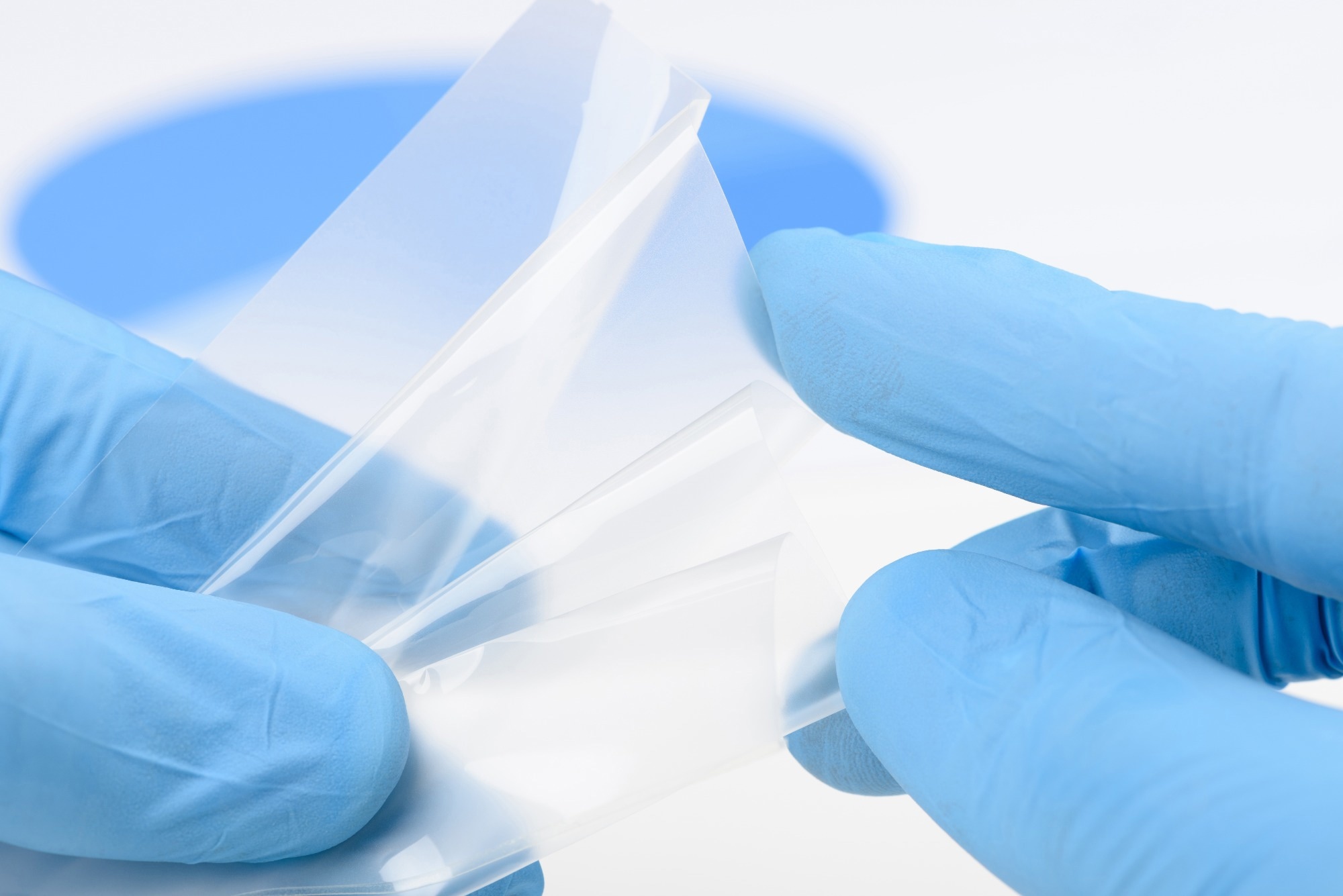 Close-up of gloved hands handling a transparent polymer film, representing advanced materials research and flexible electronic technologies